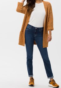 Virkat orange cardigan med vida ärmar och fickor, kombinerat med en vit T-shirt och blå jeans. Skor: svarta och orange sneakers.