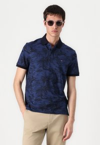 Námornícky modrá polo tričko s čiernym tropickým vzorom, krátke rukávy a čierny golier. Model má na sebe šedé slnečné okuliare a khaki nohavice.