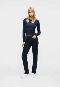 Γυναίκα όρθια φοράει στενή μπλε μπλούζα με μακριά μανίκια, σκούρο μπλε τζιν, λευκά αθλητικά παπούτσια, με το ένα χέρι στην τσέπη, μπροστά σε απλό φόντο.