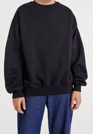 Personne portant un sweat-shirt noir ample à manches longues et un jean bleu, debout devant un fond clair uni.