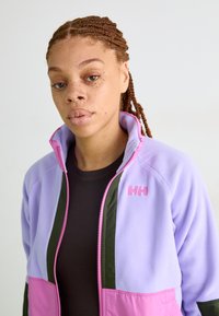 Lilla, rohelise ja roosa fleece-jakk; varustatud tõmbluku, kõrge krae ja rinna külge bordüürde logo. Pehme tekstuur.