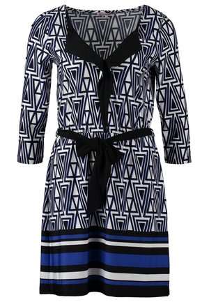 Femme portant une robe à motif géométrique noire, blanche et bleue avec des manches trois-quarts, des collants noirs, des bottines noires et un sac à main blanc.