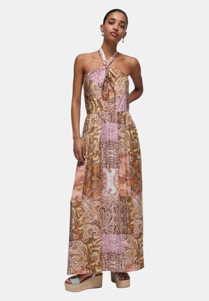 Donna che indossa un abito lungo senza maniche, con collo alla coreana, patchwork con fantasie paisley rosa, marroni e beige e sandali con zeppa.