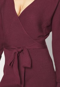 Burgundy wrapklänning i ribbat tyg, med djup V-ringning, långa ärmar och ett knytbälte i midjan.