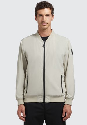 Chaqueta bomber color beige claro hecha de un tejido suave, con cremallera frontal, bolsillos laterales, puños acanalados y un logo negro en la manga.