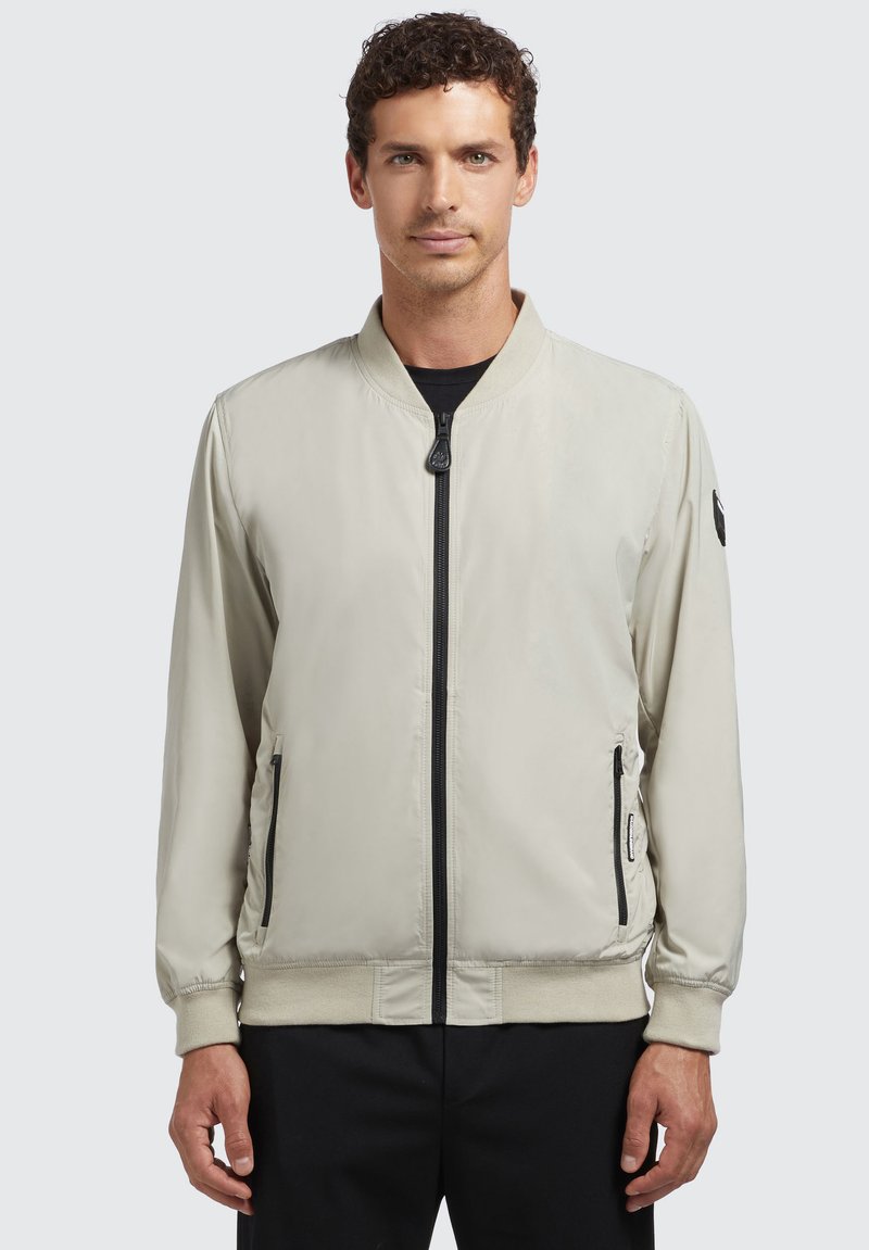 Heledad beez bomber-jakk, mis on valmistatud siledast kangast, esiküljel on lukuga, külgedel taskud, soonikkoes varrukad ja must logo varrukal.