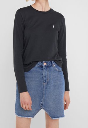 Persoon draagt een zwart shirt met lange mouwen, voorzien van een klein wit logo, en een blauwe spijkerrok met een asymmetrische, rafelige zoom.