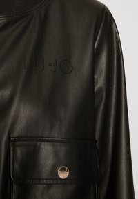 Sort læderjakke med en mock-neck, frontlomme med metallisk trykknap og perforeret "LUJO" logo på brystet.