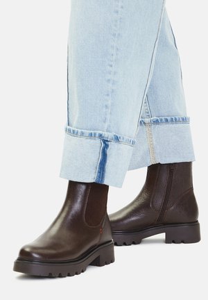 Brązowe skórzane botki za kostkę z elastycznymi panelami bocznymi, teksturowanym wykończeniem i grubą gumową podeszwą, zestawione z jasnoniebieskimi dżinsami podwiniętymi na dole.