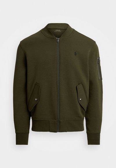 Veste bomber vert olive avec une fermeture éclair à l'avant, deux poches latérales et une poche sur la manche avec fermeture éclair. Fabriquée en matériau doux et texturé.