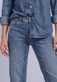 Personne portant un jean en denim bleu et une chemise en denim bleu assortie aux manches retroussées, debout, les bras détendus.