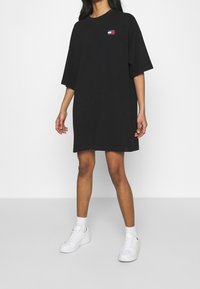Svart oversized t-shirt klänning med en liten logotyp i röd och blå på vänster bröst, tillverkad av mjukt bomullsmaterial. Buren med vita sneakers.