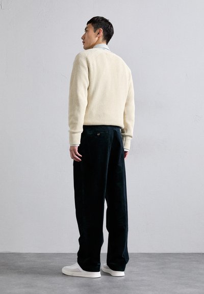 Suéter de canalé crema con un corte casual, combinado con pantalones de pana verde oscuro. El modelo lleva zapatillas blancas y está de espaldas, ligeramente alejado de la cámara.