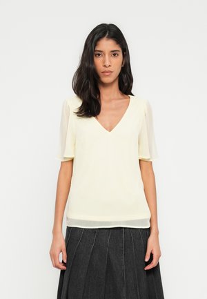 Haut à manches courtes avec col en V, en tissu jaune pâle, présentant une texture lisse et une coupe décontractée. Associé à une jupe plissée foncée.