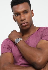 Montre rectangulaire argentée avec un écran numérique sur un bracelet en maille. Portée sur un bras musclé ; le modèle est habillé d'un t-shirt ajusté violet.