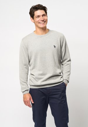 Lichtgrijze crewneck trui van een zachte breimateriaal, met ribgebreide manchetten en zoom, en een klein marineblauw logo op de borst. Draagt met donkere broek.