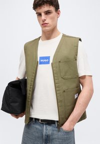 Νεαρός άνδρας που φοράει γιλέκο ελιάς πάνω από λευκό T-shirt Hugo, κρατώντας μαύρη τσάντα με σχέδια, με το ένα χέρι στην τσέπη των μπλε τζιν.