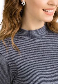 Pull gris côtelé à col montant. Le modèle porte des boucles d'oreilles circulaires en argent. Les cheveux sont ondulés et tombent sur une épaule.