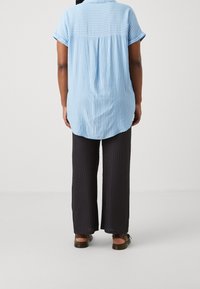 Ljust blå, randig kortärmad skjorta med bågformad nederkant, tillsammans med löst sittande, texturerade svarta byxor och bruna sandaler.