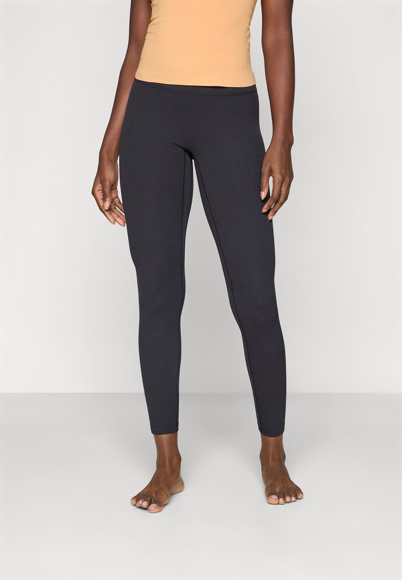 Svarte ribbede leggings med en tettsittende design, elastisk midje og ingen synlige sømmer. Båret med en hudfarget topp. Bare føtter synlige.