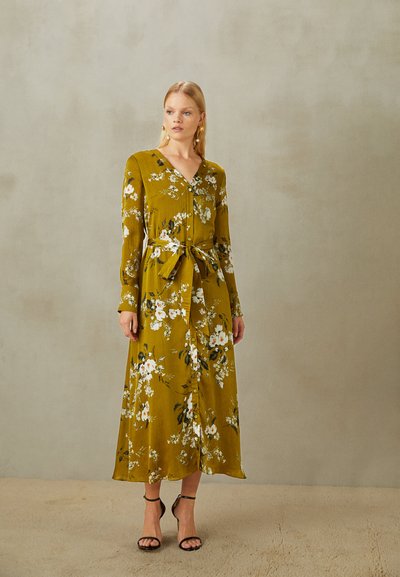 Vestido longo na cor mostarda, com padrões florais, decote em V, botões e cintura atada. Com mangas longas e uma silhueta fluida.