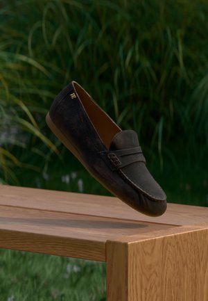 Mocassin en daim marron foncé avec une bride avant et une boucle décorative, finition légèrement texturée et bout arrondi, présenté au-dessus d'une surface en bois.