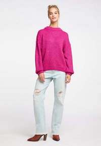 Maglione lavorato a maglia di un brillante colore rosa con un colletto alto, abbinato a jeans strappati di un leggero blu e stivaletti con tacco marroni. Texture morbida, vestibilità ampia.