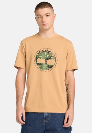 Młody mężczyzna ubrany w beżową koszulkę Timberland z zielonym logo drzewa i niebieskie dżinsy na jasnym, jednolitym tle.