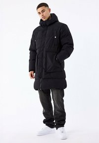 Sort pufferscoat med hætte, frontlynlås, to brystlommer og en længere skæring. Båret over mørke jeans og hvide sneakers.