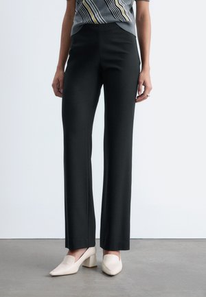 Frau trägt schwarze, weit geschnittene Hosen, gestreiftes ärmelloses Oberteil und cremefarbene Slipper mit Blockabsatz, steht auf grauem Boden vor weißer Wand.