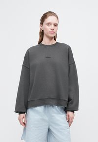 Sudadera gris oscuro con puños y dobladillo acanalados, que presenta un pequeño logotipo en el centro. Combina con pantalones cortos texturizados de color azul claro.