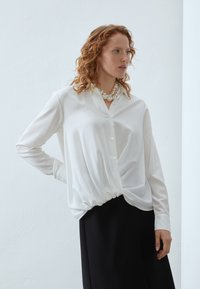 Weiße Bluse mit langen Ärmeln und Knopfleiste, mit einem gerafften Saum und einer glatten, glänzenden Textur. Kombiniert mit einem schwarzen Rock.