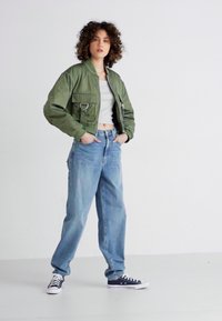 Chaqueta bomber verde ajustada con bolsillos frontales, camiseta gris ceñida y jeans holgados de color azul claro. Zapatillas de lona en blanco y negro completan el look.