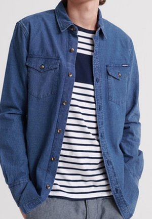 Person trägt ein aufgeknöpftes blaues Jeanshemd über einem marineblau-weiß gestreiften T-Shirt mit grauer Hose, Hände in den Taschen.