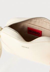 Kreemivärvi nahast käekott lukuasetusega, beež sisemus, punane silt kirjaga „HUGO” ja peene õmbluse detail välispinnal.