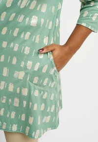 Tunic verde con un motivo a pennellate metalliche color crema, maniche lunghe e una tasca laterale con una superficie liscia.