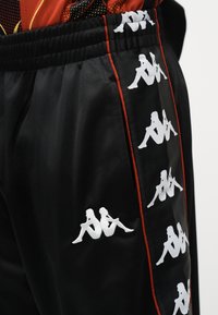 Μαύρο αθλητικό παντελόνι με λευκά λογότυπα Kappa κατά μήκος της πλευρικής ρίγας, περιτριγυρισμένη από λεπτή κόκκινη ταινία.