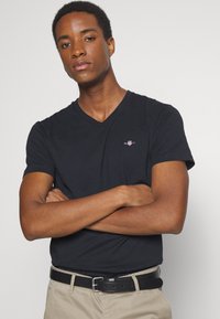 Camiseta negra de cuello en V hecha de algodón, de manga corta, con un pequeño logo en el pecho. Lleva un cinturón negro y pantalones beiges.