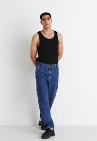Black sleeveless tank top with blue denim jeans. Jeans feature a relaxed fit and front pockets. Model wears black shoes. Simple, minimal design.
