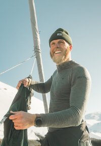 Mies, joka halaa partaa ja käyttää pipolakkia, säätää köyttä purjeveneessä, pukeutuneena pitkähihaiseen paitaan ja kelloon, taustalla lumiset vuoret ja kirkas taivas.