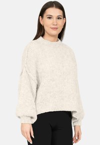 Jonge vrouw met stijlbruin haar, goudkleurige oorringen, een los witte gebreide trui en zwarte broek, staand tegen een witte achtergrond.