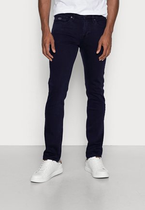 Mann trägt dunkelblaue Slim-Fit-Jeans und weiße Sneaker, steht vor einem schlichten hellen Hintergrund.