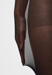 Gros plan sur une main portant des collants transparents foncés, avec les doigts légèrement courbés et les bouts des ongles visibles sur un fond clair.