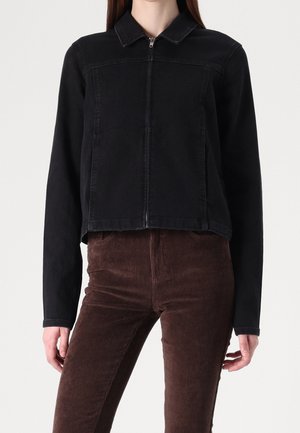 Kobieta ubrana w czarną kurtkę na zamek z długimi rękawami i brązowe spodnie sztruksowe, stoi na tle jednolitego jasnego tła.