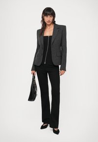 Femme portant un blazer à rayures noires et grises, un haut zippé noir, un pantalon noir, des chaussures noires à bout pointu, tenant un sac à main noir brillant.