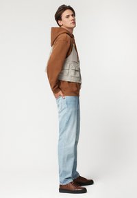 Brązowa bluza z kapturem noszona pod szarym kamizelką z wieloma kieszeniami, w połączeniu z jasnoniebieskimi dżinsami i brązowymi trampkami. Gładki materiał i luźny krój.