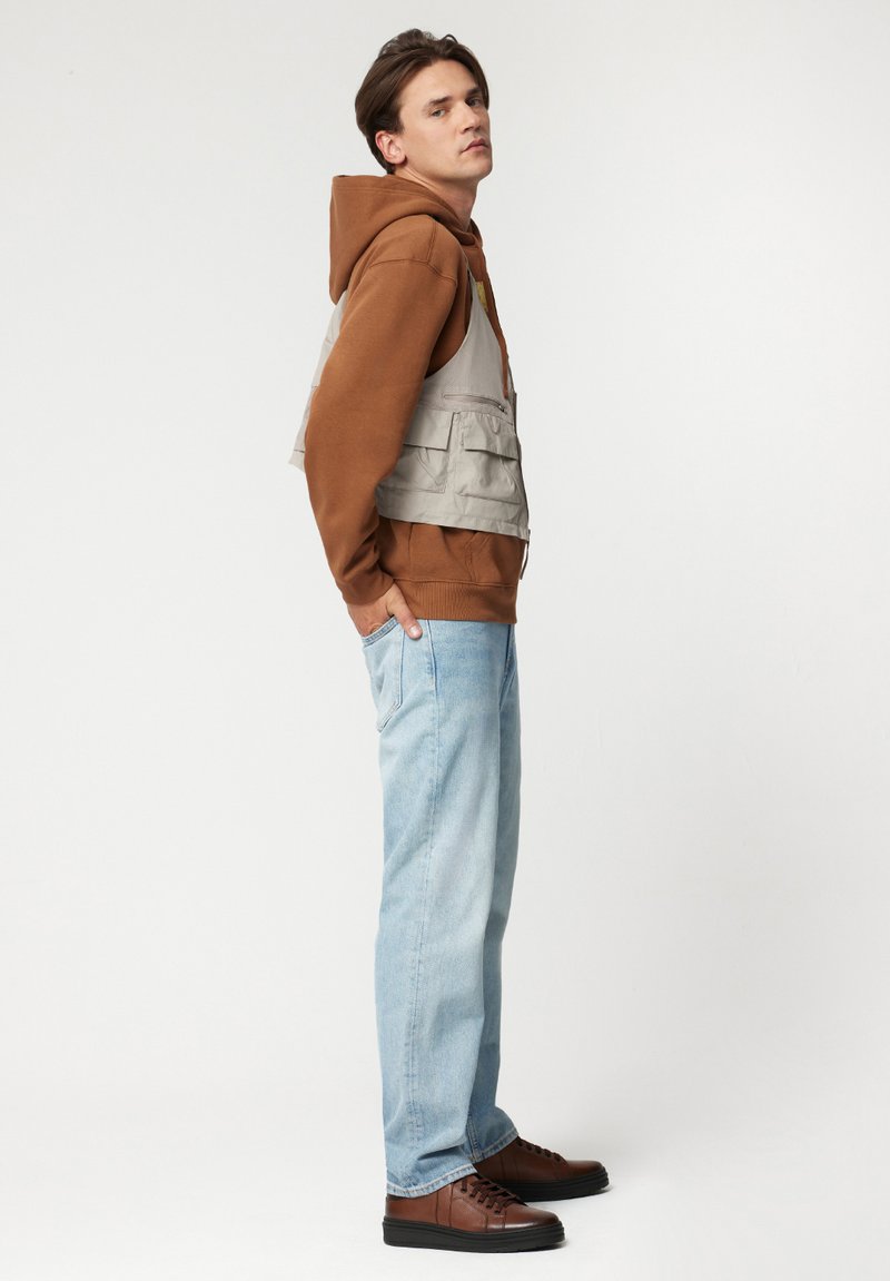 Brązowa bluza z kapturem noszona pod szarym kamizelką z wieloma kieszeniami, w połączeniu z jasnoniebieskimi dżinsami i brązowymi trampkami. Gładki materiał i luźny krój.