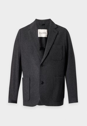 Joven con blazer gris oscuro, camisa negra, vaqueros azul claro y mocasines negros, de pie con una mano en el bolsillo contra un fondo blanco.