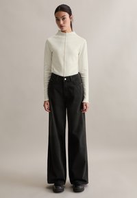 Ecru bluzka z długim rękawem i ozdobnym dekoltem w kształcie fali, zestawiona z czarnymi, szerokimi spodniami z wysokim stanem. Gładka faktura i dopasowany krój.