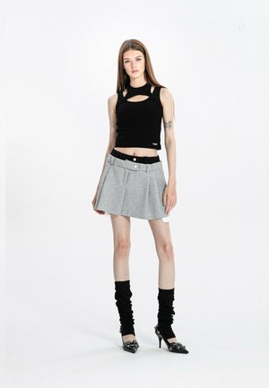 Joven mujer con un top negro sin mangas, minifalda gris plisada, calentadores de piernas negros y zapatos de tacón negro, de pie contra un fondo blanco.